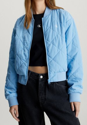 Kvinde iført lyseblå quiltet bomberjakke over sort cropped Calvin Klein top og højtaljede sorte jeans, står med afslappet kropsholdning.