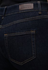 Σκούρο μπλε τζιν από denim με πορτοκαλί ραφές, που δείχνουν την πίσω τσέπη και μια καφέ δερμάτινη ετικέτα επωνυμίας που γράφει "ONLY."