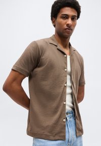 Mann iført en brun kortermet skjorte med knapper over en hvit tanktopp og lyseblå jeans, stående mot en ensfarget lys bakgrunn.