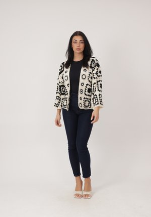 Donna in piedi che indossa una maglia nera, un cardigan con motivo bianco e nero, jeans scuri e sandali bianchi aperti con tacco su sfondo neutro.