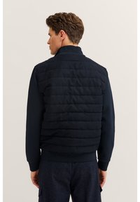 Navy blue puffer vest with quilted design, featuring a high collar and elastic fitted hem, worn over a long-sleeve shirt.