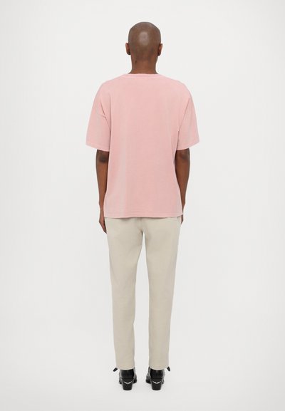 T-shirt en coton rose clair avec une coupe décontractée, des manches courtes et des détails de couture visibles, associé à un pantalon beige fuselé et des chaussures à talons noires.