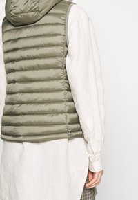 Hedvábně lesklá olivově zelená vesta s kapucí, vyrobená z hladkého materiálu. Má vertikální švy a nosí se přes světlou košili.