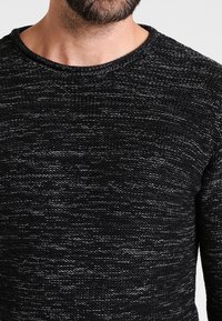 Pull noir en maille avec un motif texturé, un col rond et une coupe décontractée. Le tissu a une texture douce et légèrement irrégulière.