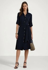 Žena v námořnicky modrých košilových šatech s knoflíky a s ohrnutými rukávy, černých lodičkách na kotníkovém pásku, náramcích a držící pruhovanou tkanou kabelku.