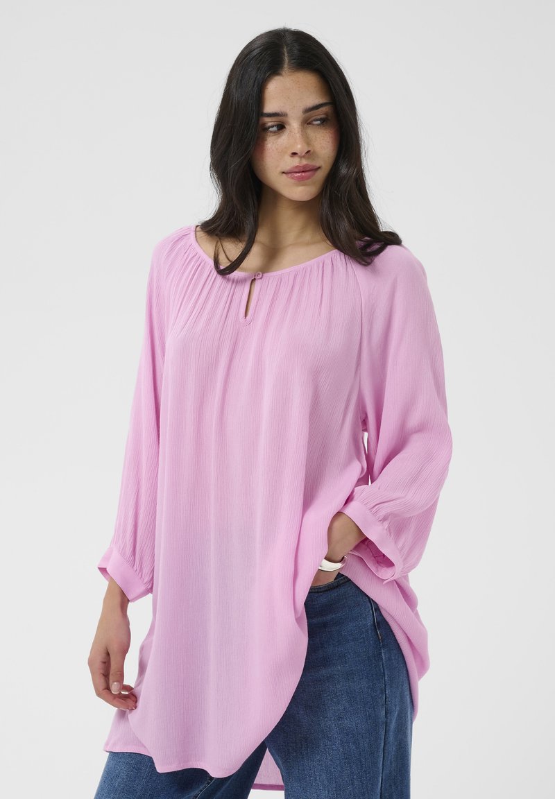 Femme portant un chemisier rose ample à manches longues et un jean bleu, une main dans la poche, regardant légèrement vers sa droite.