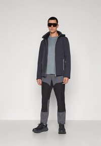 Jachetă cu glugă închisă la culoare navy, cămașă albastru deschis și pantaloni gri-negru, cu model. Adidași cu tălpi texturate. Stil casual de exterior, cu detalii hardware minimaliste.