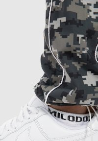 Camouflage-mønstrede joggingbukser i nuancer af grå og sort med hvide detaljer, parret med hvide sneakers med et sort logo på sokken.