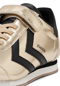 Goldfarbene Metallic-Sneaker mit schwarzen Akzenten, strukturiertem Obermaterial, flachen Schnürsenkeln und einem Klettverschluss. Das "STADION"-Logo ist an der Seite sichtbar.