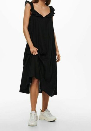 Mujer con un vestido midi negro sin mangas con tirantes con volantes y zapatillas blancas gruesas, de pie frente a un fondo blanco.