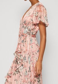 Blomsterindpakket kjole i lys pink med løse flæseærmer, dyb V-hals og volangkant. Har en belagt talje og lodrette striber.