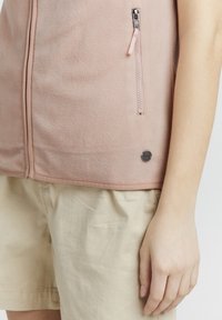 Ljusrosa fleeceväst med dragkedja fram, sidoficka och rund logotypdetalj; matchad med ljusbeiga shorts. Mjuk textur.