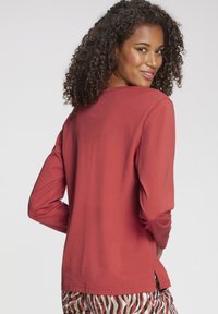 Rotes langärmliges Shirt aus weichem Stoff, mit rundem Ausschnitt und seitlichen Schlitzen, kombiniert mit gemusterten Hosen.