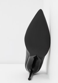 Chaussure à talon stiletto noire à bout pointu reposant sur le côté contre une surface blanche et un mur.