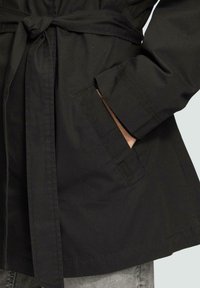 Schwarze, textile Oberbekleidung mit einem Gürtel, die eine glatte Textur, einen geraden Schnitt und seitliche Taschen aufweist. Sichtbare Nähdetails an den Ärmeln.