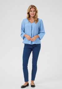 Blouse bleue claire à manches longues avec un col en V, légèrement évasée en bas, et une finition texturée, associée à un jean skinny noir et des ballerines noires.