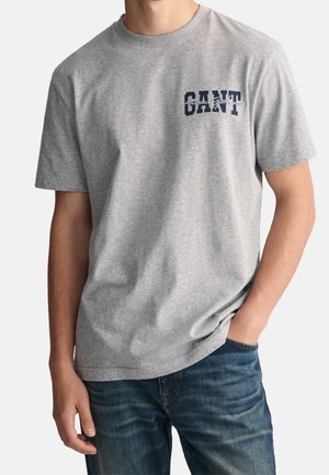 Mężczyzna w jasnoszarej krótkiej koszulce GANT z granatowym napisem i niebieskich dżinsach, z prawą ręką w kieszeni, na jednolitym tle.
