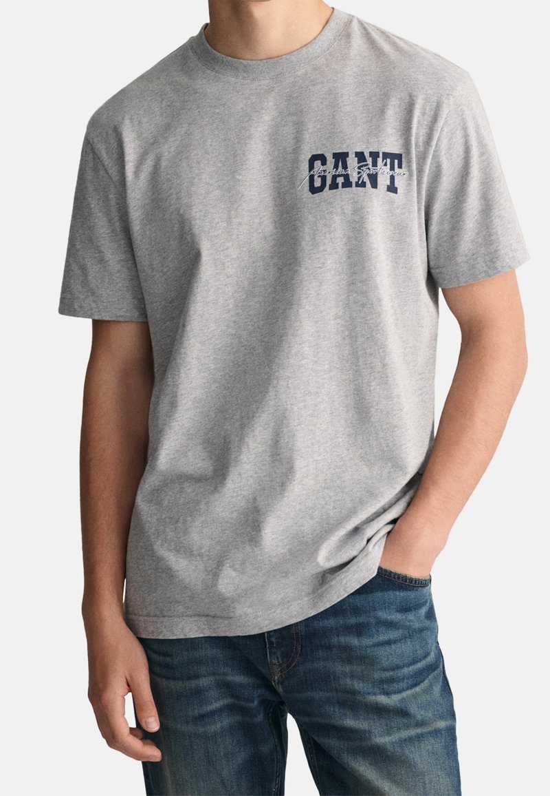 Homme portant un t-shirt GANT gris clair à manches courtes avec texte bleu marine et un jean bleu, main droite dans la poche, sur fond uni.