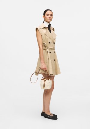 Woman wearing sleeveless beige trench dress with black buttons, holding small white quilted handbag, and black flat shoes.