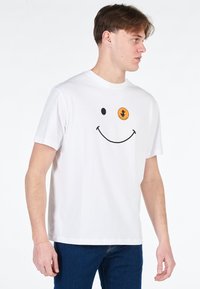 T-shirt bianca in cotone con una grafica di faccina sorridente nera e un accento arancione sul petto. Maniche corte e vestibilità rilassata.