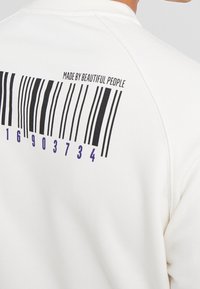 Sudadera blanca con un gráfico de código de barras negro en la parte de atrás, con acentos morados y texto que dice "HECHO POR GENTE BELLA".