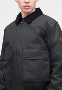 Homme portant une veste noire zippée avec de grandes poches avant et un col en velours côtelé, visible de la moitié de la poitrine jusqu'au menton.