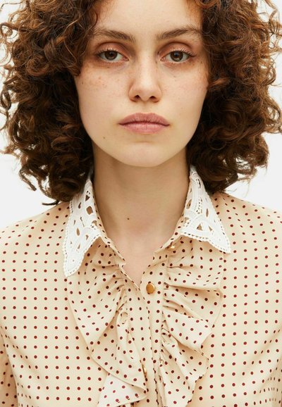 Beige blouse met stippen, voorzien van een geplooide voorkant, kanten kraagdetails en gouden knopen. De stof lijkt lichtgewicht en textuur te hebben.