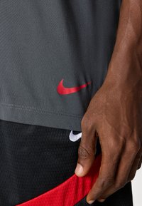 Pilkai sportsiniai marškiniai, pagaminti iš lygaus audinio, su raudona Nike logo apacioje. Juodos šortai su tekstūruotu raštu ir matoma raudona juosta.