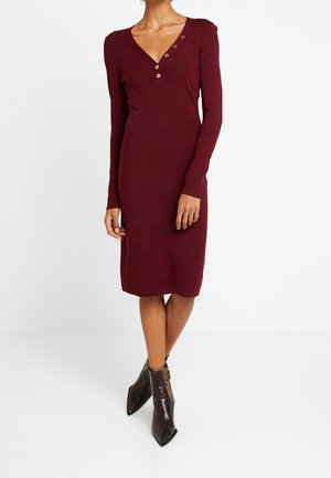 Femme portant une robe midi ajustée à manches longues bordeaux avec un col en V à trois boutons et des bottines marron à bout pointu avec un motif texturé.
