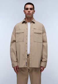 Camisa de algodón beige con un corte relajado, dos bolsillos frontales y un ligero brillo. Presenta un cuello con solapa y cierres de botón.