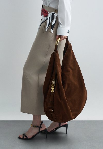 Γυναίκα με μπεζ φούστα και λευκό πουκάμισο να κρατά μεγάλη καφέ σουέντ τσάντα Moschino με χρυσή λαβή, φορώντας μαύρες κοτλέ επιλογές.