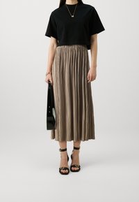Svart cropped t-shirt, plisserad beige kjol, svarta strap-heeled skor, håller en glansig svart väska. Kjolen har vertikala plisseringar och elastisk midja.