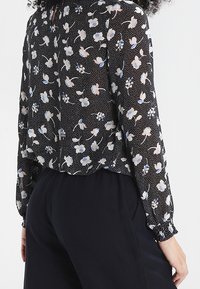 Czarna, przezroczysta bluzka z kwiatowym wzorem w niebiesko-białe kolory, z okrągłym dekoltem, długimi rękawami i marszczonymi mankietami.