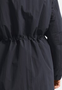 Donkerblauwe windbreaker met gathered taille detail, gladde textuur, lange mouwen en elastische manchetten. Lichtgewicht en waterafstotende stof.