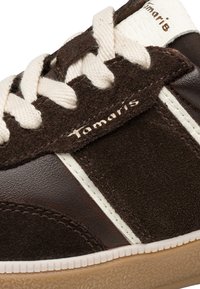 Gros plan sur une basket en daim et cuir marron et blanc avec des lacets blancs et le logo "Tamaris" sur le côté et la languette.