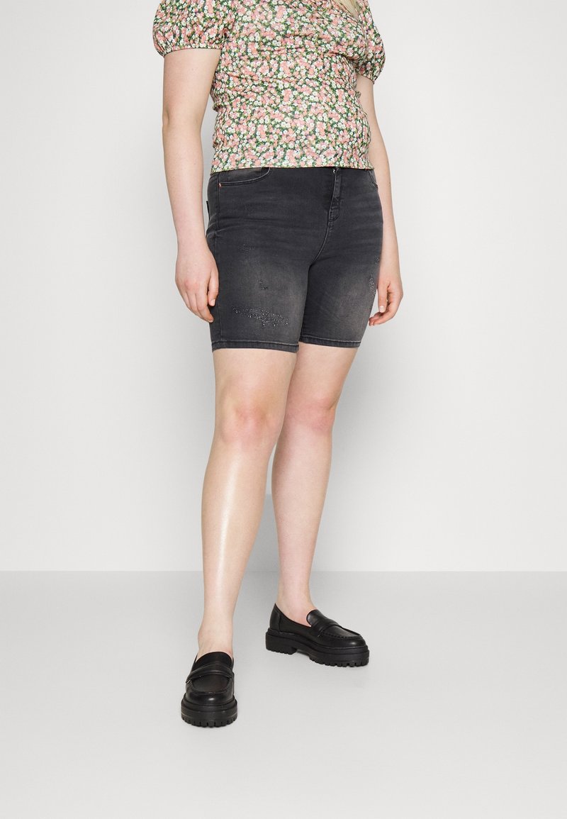 Black denim Bermuda shorts with light distressing, paired with a floral patterned short-sleeve top. Person wears black loafers.