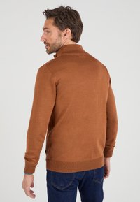 Suéter marrón de punto con cuello alto, mangas largas, puños y dobladillo acanalados. Presenta una abertura lateral y una textura sutil. Se combina con jeans oscuros.