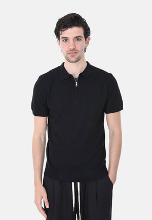 Giovane uomo con capelli scuri e ricci, che indossa una polo nera a maniche corte in maglia e pantaloni neri con coulisse bianche, su uno sfondo semplice.