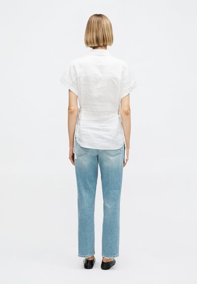 Άτομο με κοντά ξανθά μαλλιά φοράει λευκό κοντομάνικο πουκάμισο, ανοιχτό μπλε ίσια τζιν και μαύρα παπούτσια χωρίς κορδόνια, στέκεται μπροστά σε λευκό φόντο.