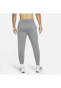 Mann in hellgrauen Jogginghosen und weißen Nike-Sneakers, barfuß stehend von hinten vor einem weißen Hintergrund.