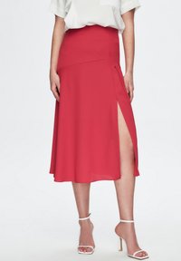 Falda midi roja con una abertura alta, confeccionada en tejido ligero. Incluye una cinturilla ajustada y un diseño fluido. Combinada con tacones blancos.