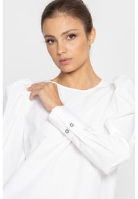 Biała bluzka z bufkami, gładki materiał bawełniany, okrągły dekolt i mankiety zapinane na guziki z kontrastowymi czarnymi guzikami.