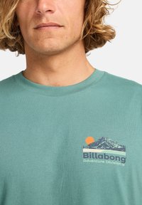 Grøn bomulds T-shirt med rund halsudskæring, prydet med et grafisk motiv af bjerge og en sol i orange og blå, samt teksten "Billabong Adventure Division".