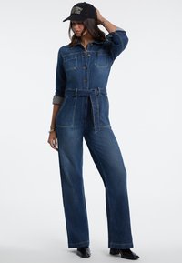 Donna che indossa una tuta di jeans blu scuro con cintura, maniche arrotolate, stivali neri e berretto nero, in posa con una mano sulla testa.