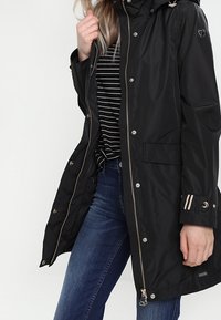 Parkas impermeabile nero con cappuccio, chiusura con zip, dettagli con bottoni e polsini a righe. Presenta due tasche frontali.