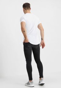 Camiseta blanca de algodón, jeans ajustados negros y zapatillas deportivas grises. Vista desde atrás, mostrando una silueta ajustada y un diseño sencillo.