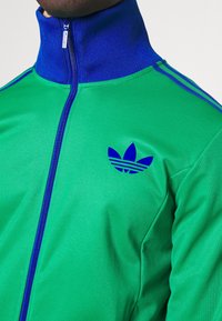Vihreä vetoketjullinen urheilutakki, jossa on sininen kaulus ja kolmen raidan koristeet. Vasemmassa rinnassa sininen Adidas-logo. Tasainen kangaspinta.