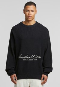 Pullover in maglia nera con una texture a coste, svasato, e logo ricamato che recita "Another Cotton Established 2023" in bianco.