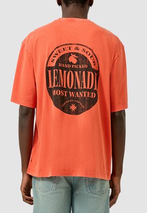 Personne portant un T-shirt orange corail avec un graphique circulaire noir "Sweet & Sour Hand Picked Lemonade Most Wanted" dans le dos.