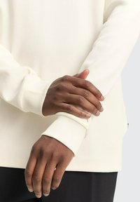 Creme lange mouwen geribbelde shirt met aansluitende manchetten, met een zachte textuur en een relaxte pasvorm. Handen gekruist over de polsen, met huidskleur zichtbaar.